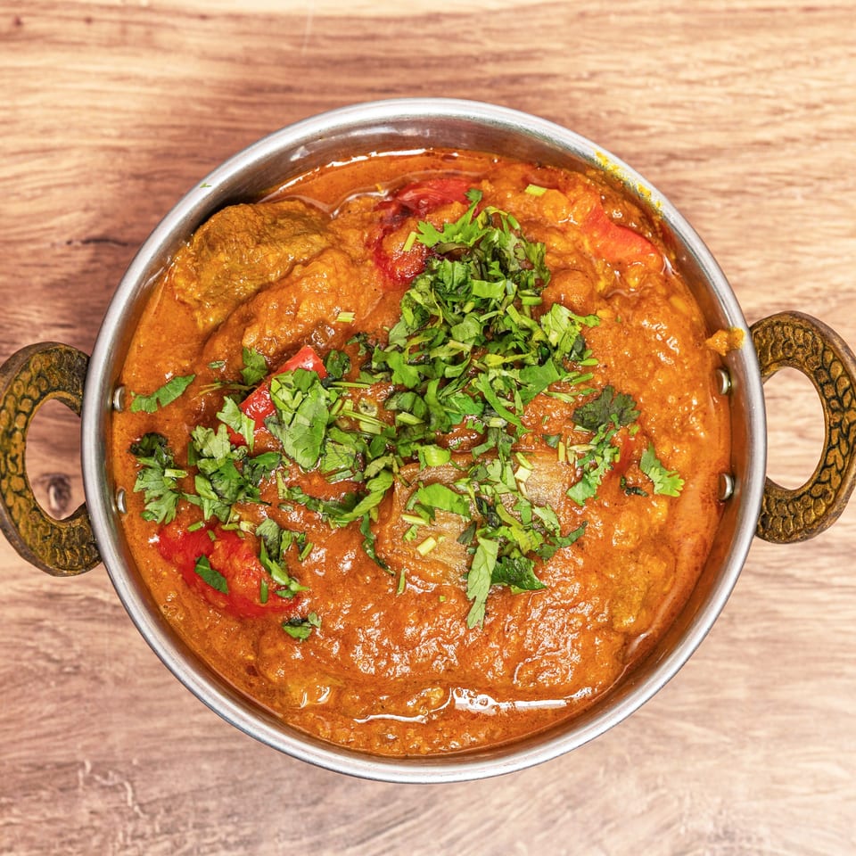 Overhead view of rich Indian curry with vegetables and fresh cilantro in a handled metal serving bowl on a wooden table.