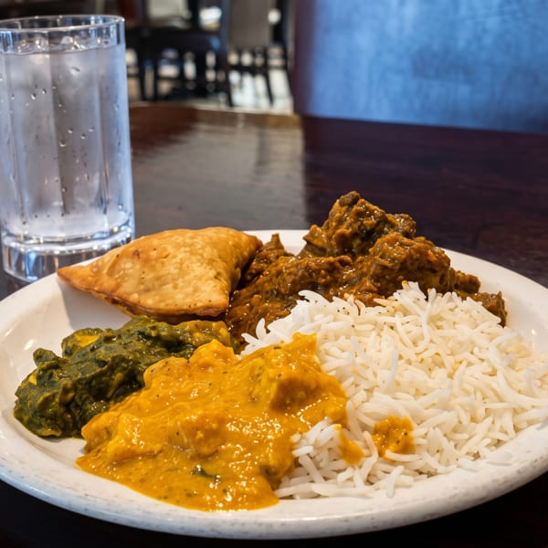 A white plate with Indian food, including rice, curries, and a samosa, next to a glass of water.