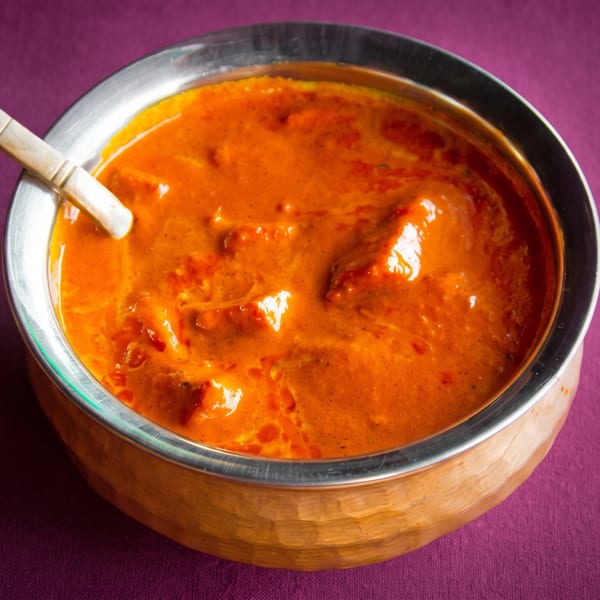 A copper bowl filled with vibrant orange butter chicken, with a wooden spoon resting inside.