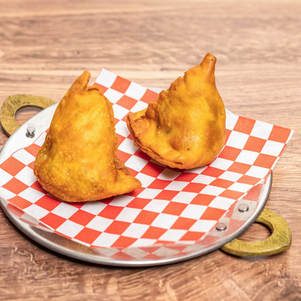 Three golden fried samosas on red-and-white checkered paper atop a metal serving tray on a wooden table.