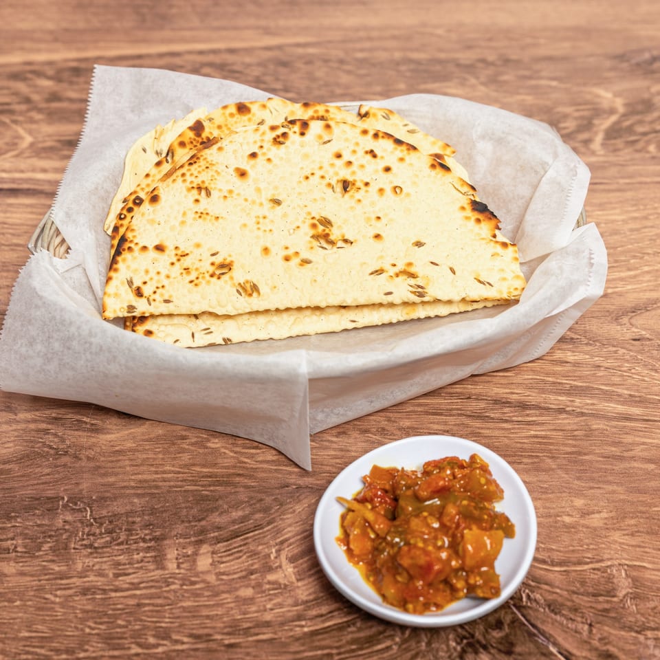 Stack of toasted, cumin-speckled flatbreads in a paper-lined basket with spicy vegetable relish on a wooden table.