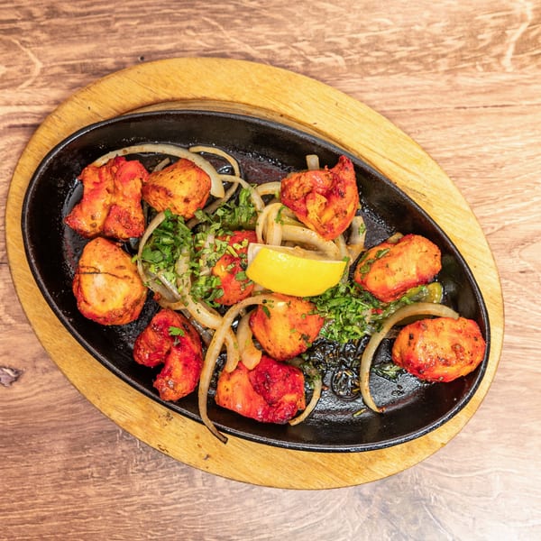 Overhead view of sizzling tandoori chicken with onions, cilantro, and lemon in a black skillet on a wooden board.