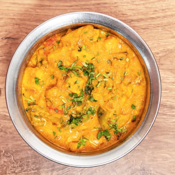Overhead view of creamy orange curry with chunks of paneer, tomatoes, and cilantro in a metal bowl on a wooden table.