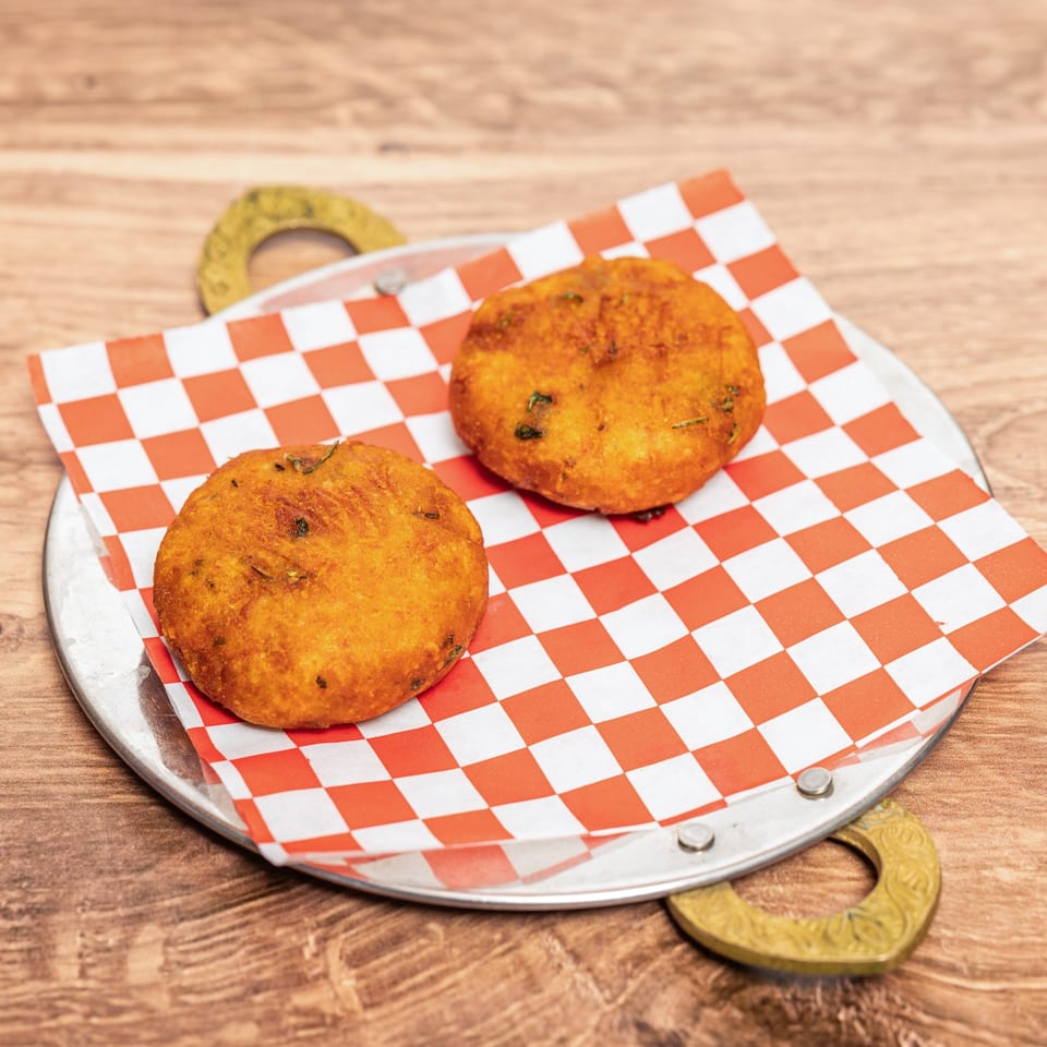 Two golden, herb-speckled fried patties sit on red-and-white checkered paper atop a metal serving tray.