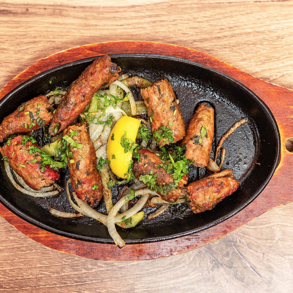 Grilled spiced meat kebabs garnished with cilantro, served with sautéed onions, peppers, and lemon in a cast-iron platter.
