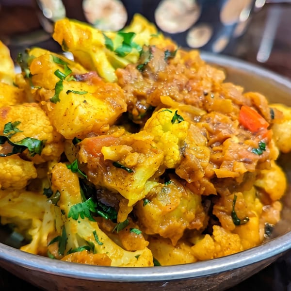 Close-up of a metal bowl filled with vibrant yellow cauliflower and potato curry, garnished with fresh cilantro.
