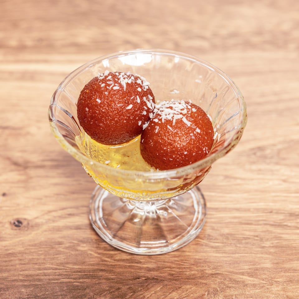 Two syrup-soaked brown dessert balls topped with shredded coconut in a glass bowl on a wooden table.