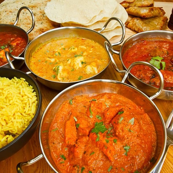 Four bowls of Indian curries, yellow rice, naan bread, and samosas on a wooden table.