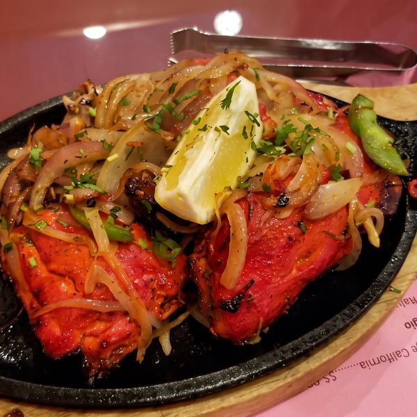 Sizzling red tandoori chicken with onions, peppers, and a lemon wedge on a black skillet.