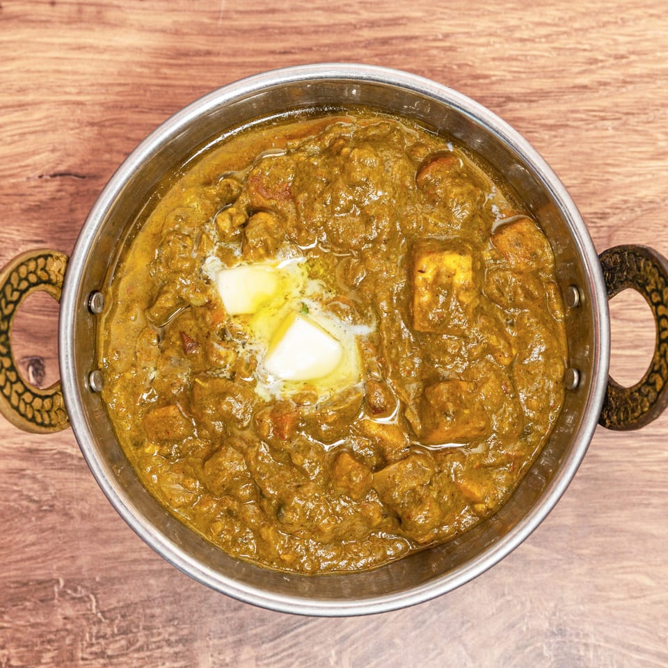 Overhead view of creamy Indian curry with paneer pieces and butter in a handled metal serving pan on a wooden table.