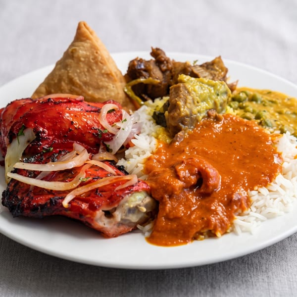 A white plate with a colorful Indian meal including tandoori chicken, rice, samosa, and curries.