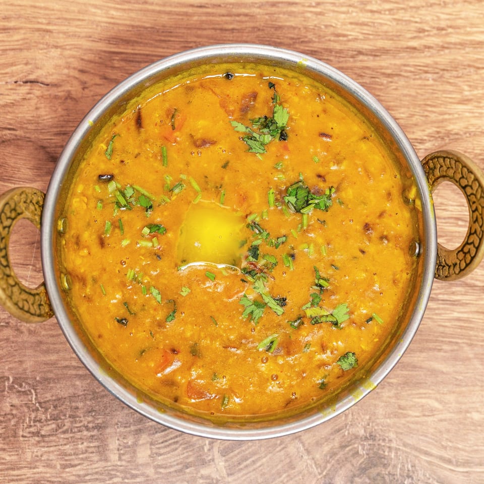 Top-down view of a golden Indian curry garnished with fresh cilantro in a handled metal serving dish on a wooden table.