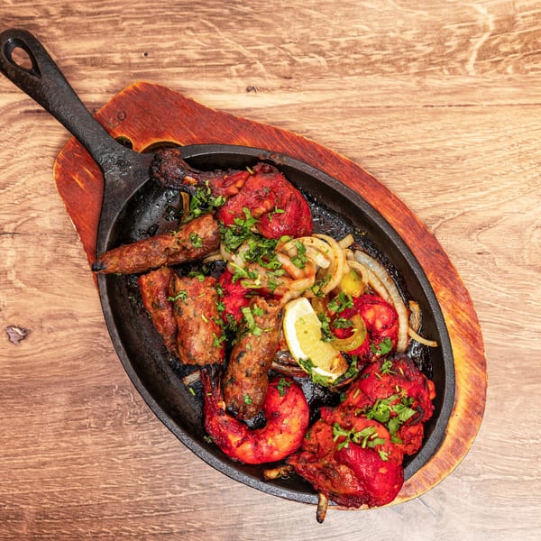 Assorted grilled kebabs, red-marinated chicken, vegetables, onions, lemon, and herbs served in a cast-iron skillet on a wooden board.