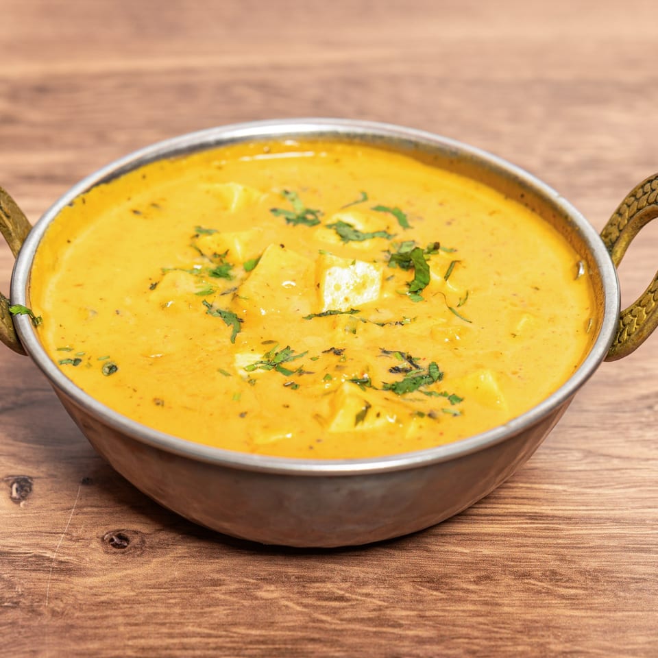 Creamy yellow paneer curry garnished with cilantro, served in a decorative metal bowl on a wooden table.